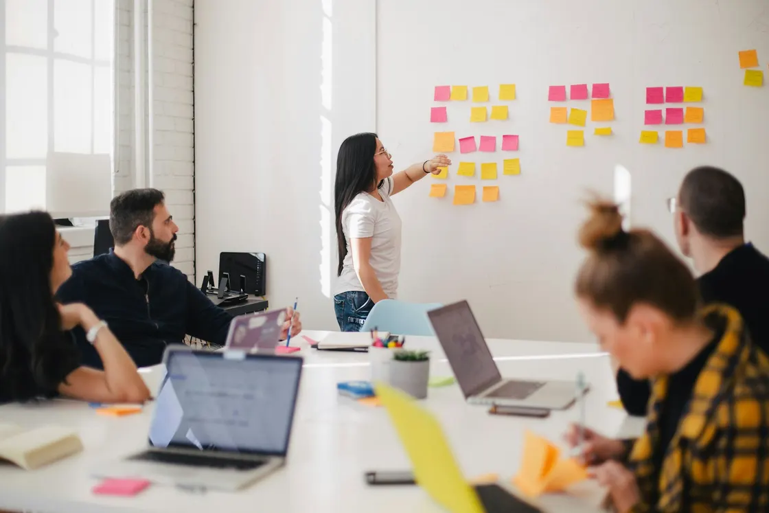 Team member pointing to sticky notes on a wall during a planning session with colleagues at laptops