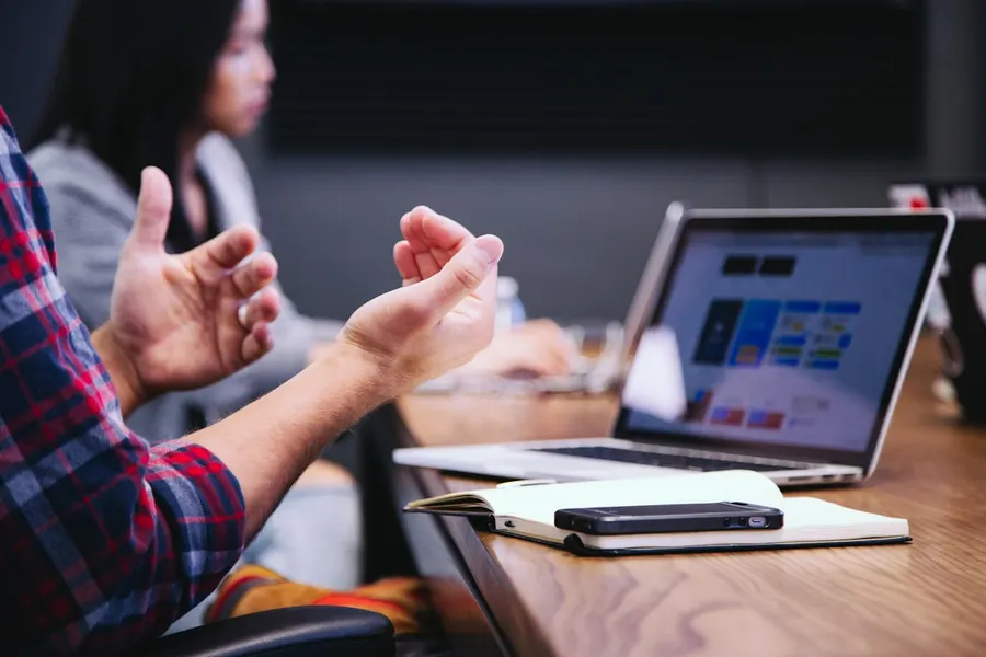 Person gesturing while explaining an idea in a meeting, laptop open on the table