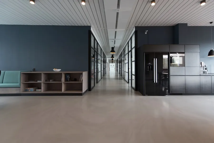 Empty modern office corridor lined with glass-walled rooms beside a dark kitchen area